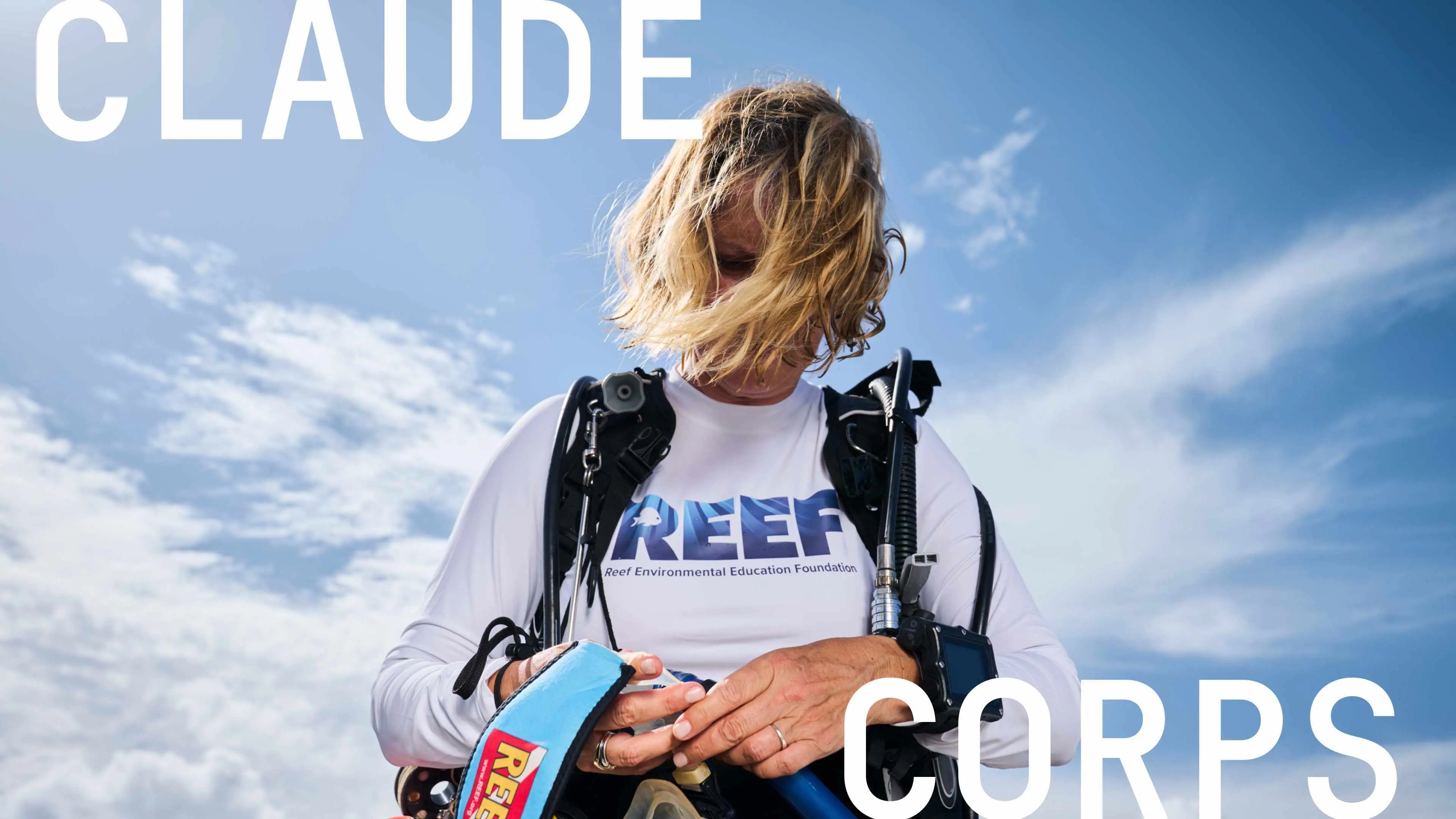 A diver in a long-sleeve white REEF shirt adjusts scuba gear against a bright blue, cloud-streaked sky.